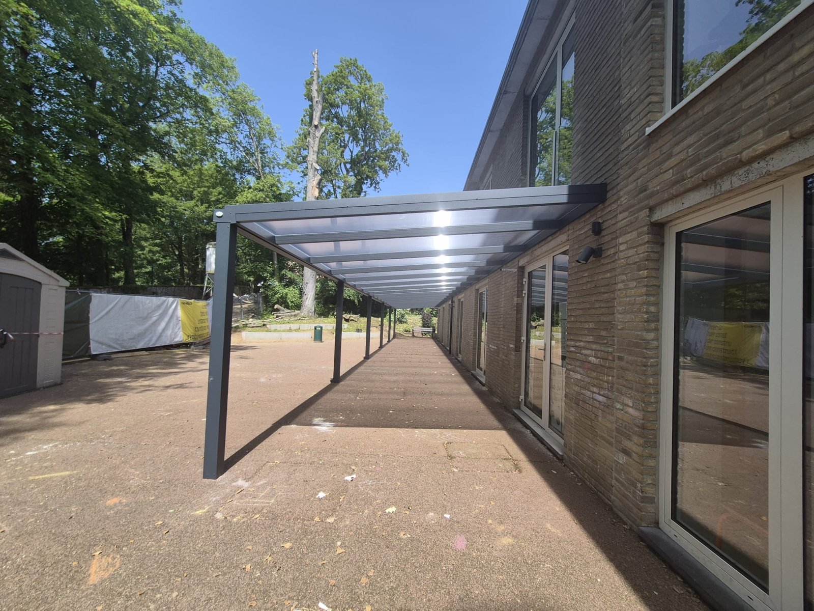 Overkapping met plat dak langs een schoolgevel, aluminium kolommen en doorschijnend dak